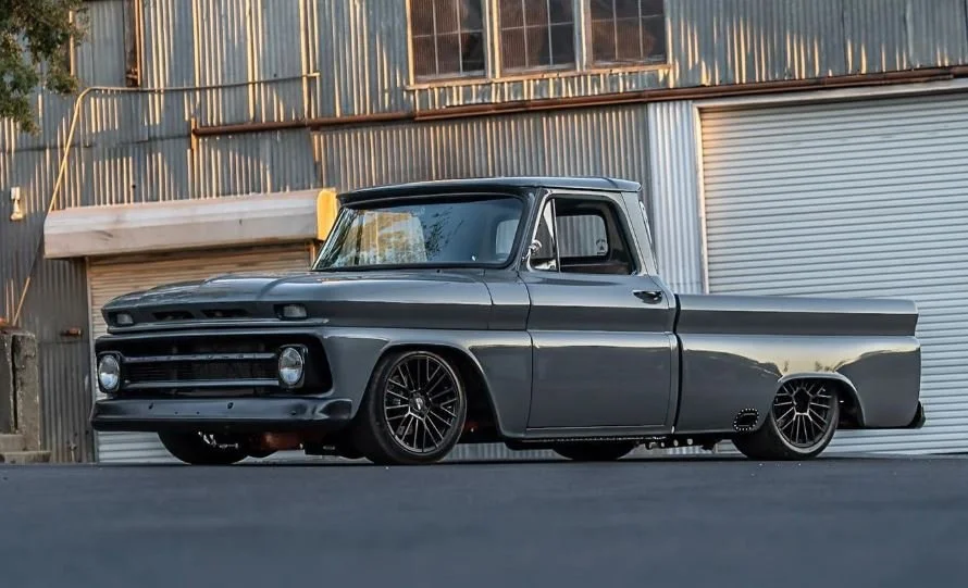A customized vintage pickup truck with a matte gray finish parked in front of a metal industrial building.