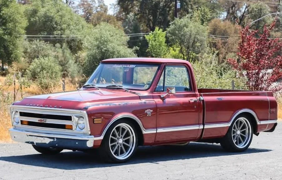 A vintage red Chevrolet pickup truck parked outdoors on a sunny day with green trees and shrubs in the background.