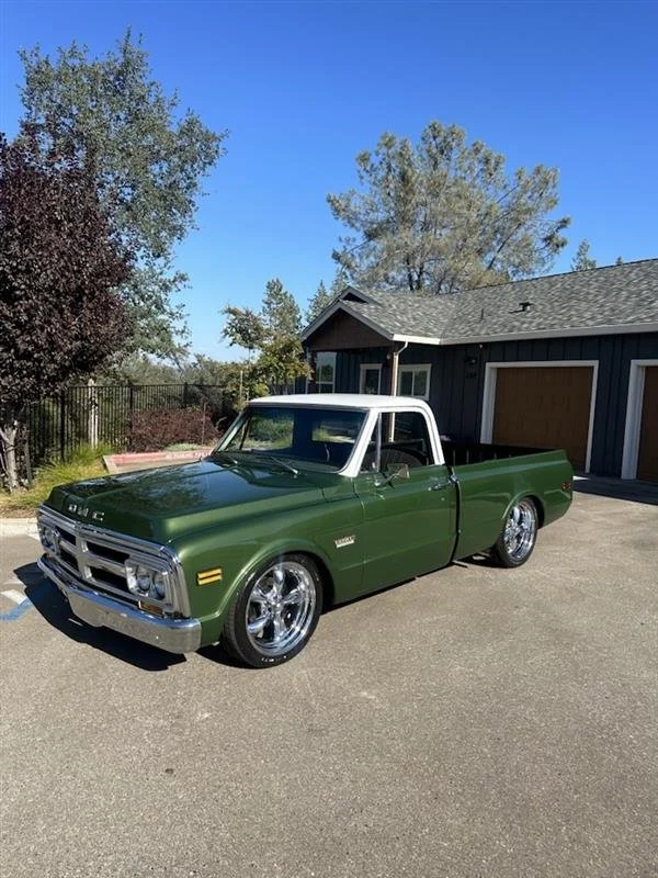 A vintage green pickup truck with a white roof parked on a driveway in front of a blue house with a garage. Trees and a clear blue sky are visible in the background.