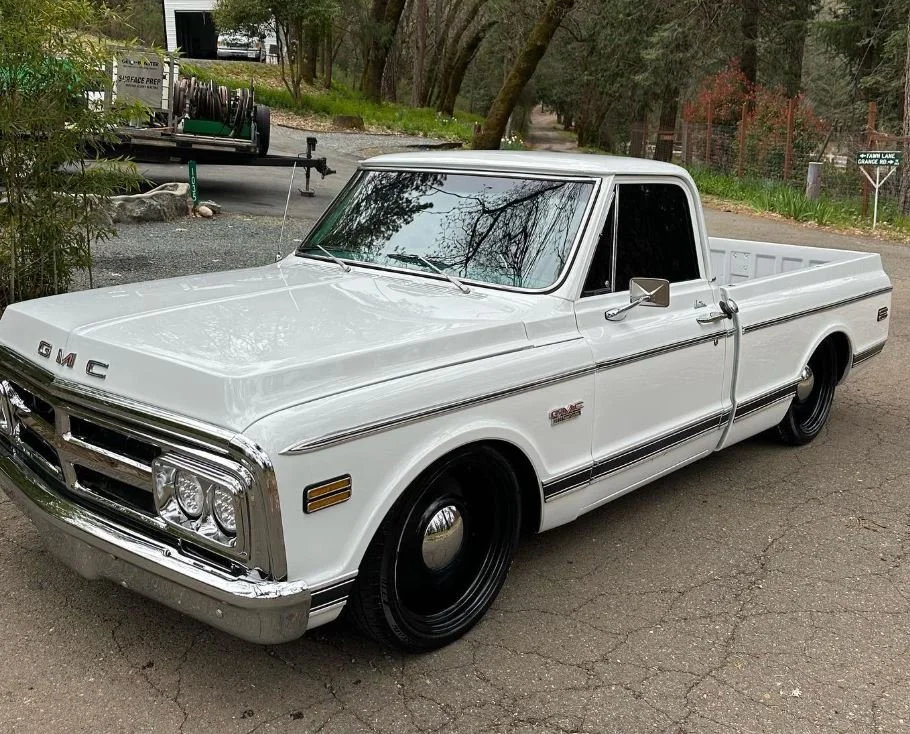 A white vintage GMC pickup truck parked on a gravel surface surrounded by trees and greenery.