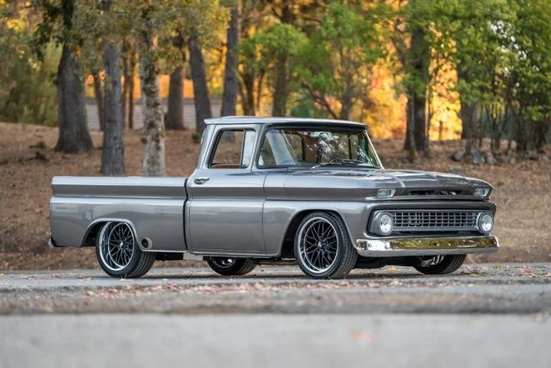 A vintage silver pickup truck with black custom wheels parked on the side of a road, with autumn trees in the background.