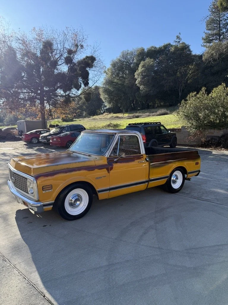A vintage Chevrolet pickup truck painted in yellow with purple accents parked in a lot during daytime, surrounded by trees and other vehicles.