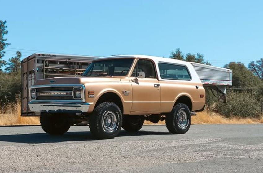 A tan vintage SUV with a cargo trailer attached, parked on a gravel road with a clear blue sky and trees in the background.
