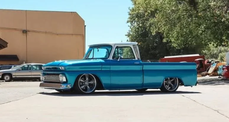 Classic vintage blue pickup truck parked outdoors with other vehicles and green trees in the background.