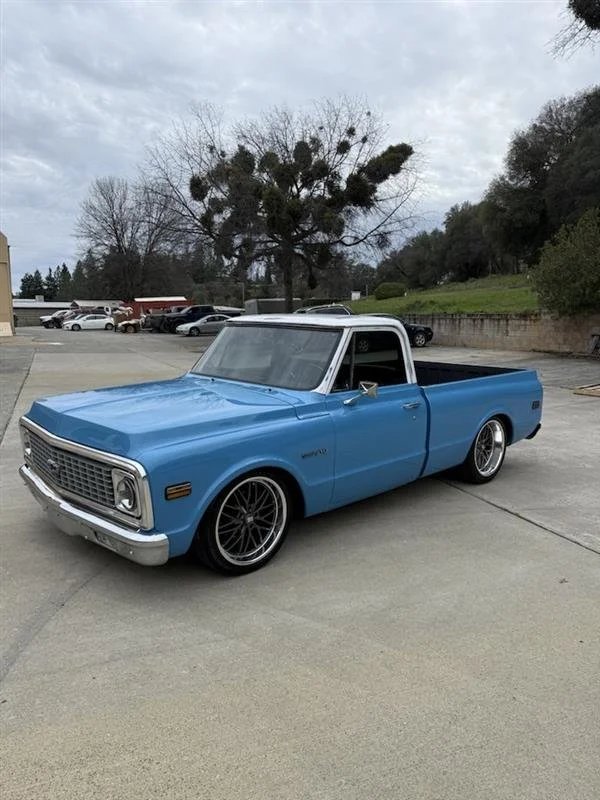 A vintage blue Chevrolet pickup truck parked in an outdoor lot with trees and cloudy sky in the background.