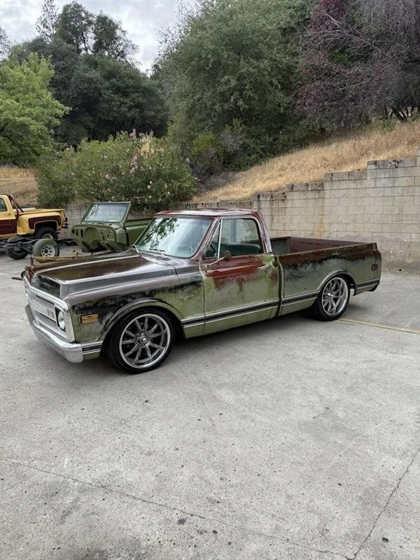 A rusty, old pickup truck with a lowered stance and aftermarket wheels parked on a driveway. There are other vintage vehicles and trees in the background.