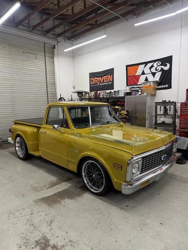 A vintage yellow Chevrolet pickup truck parked inside a workshop with tools and car parts on shelves. There are banners on the wall, including one for K&N and Driven Racing Oil.