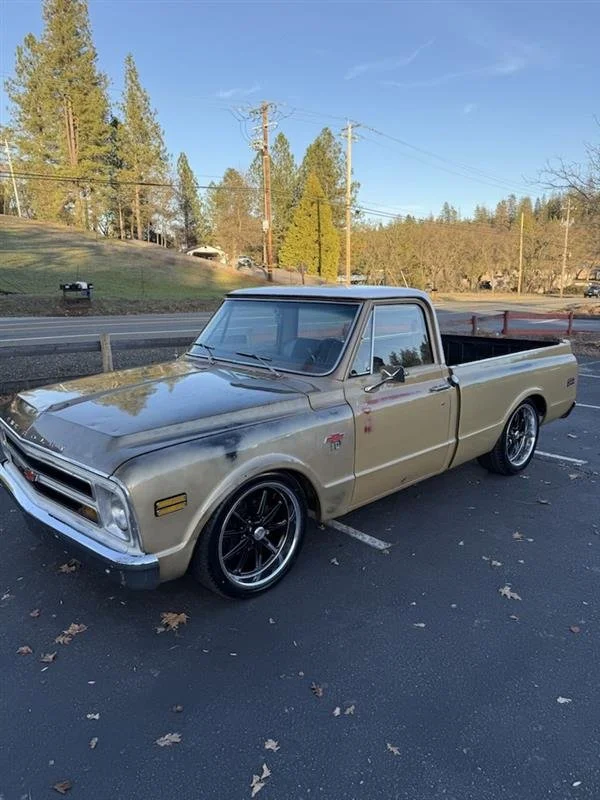 A vintage beige pickup truck with black custom wheels parked in a parking lot near trees and power lines.