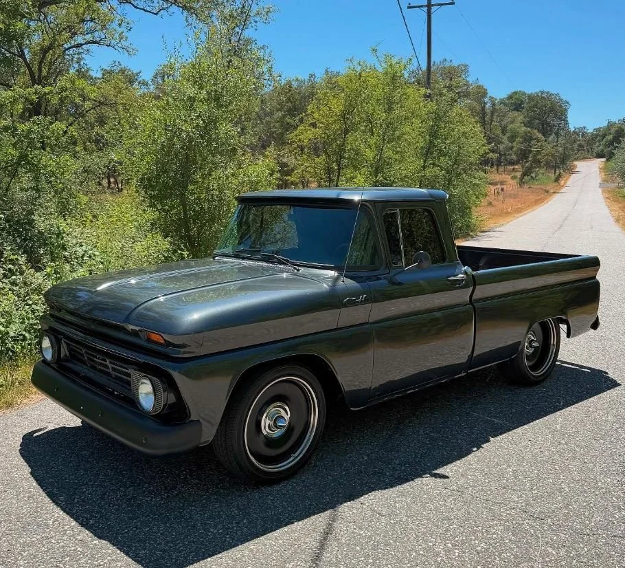 A vintage black pickup truck parked on a country road with trees and utility poles, under a clear blue sky.
