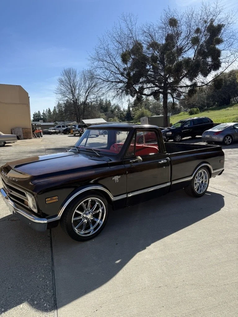 A black vintage pickup truck parked outdoors on a sunny day, with a tree and several other vehicles in the background.