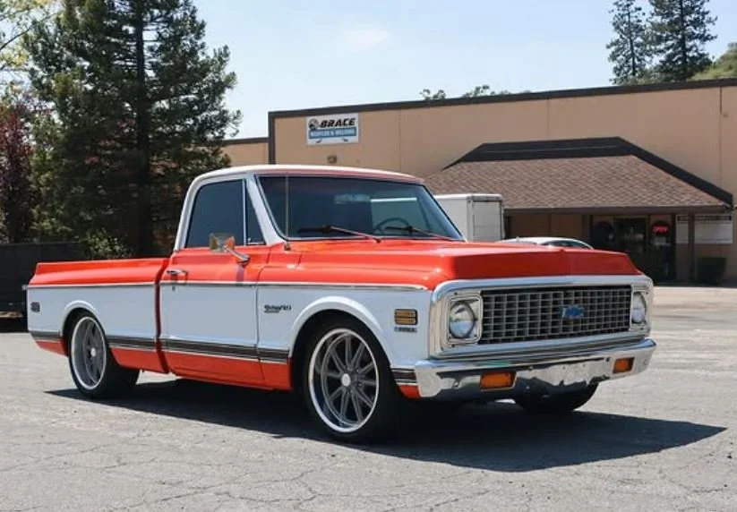 A vintage two-tone pickup truck with a red and white paint job, custom wheels, and lowered suspension parked on a paved lot in front of a building with trees in the background.