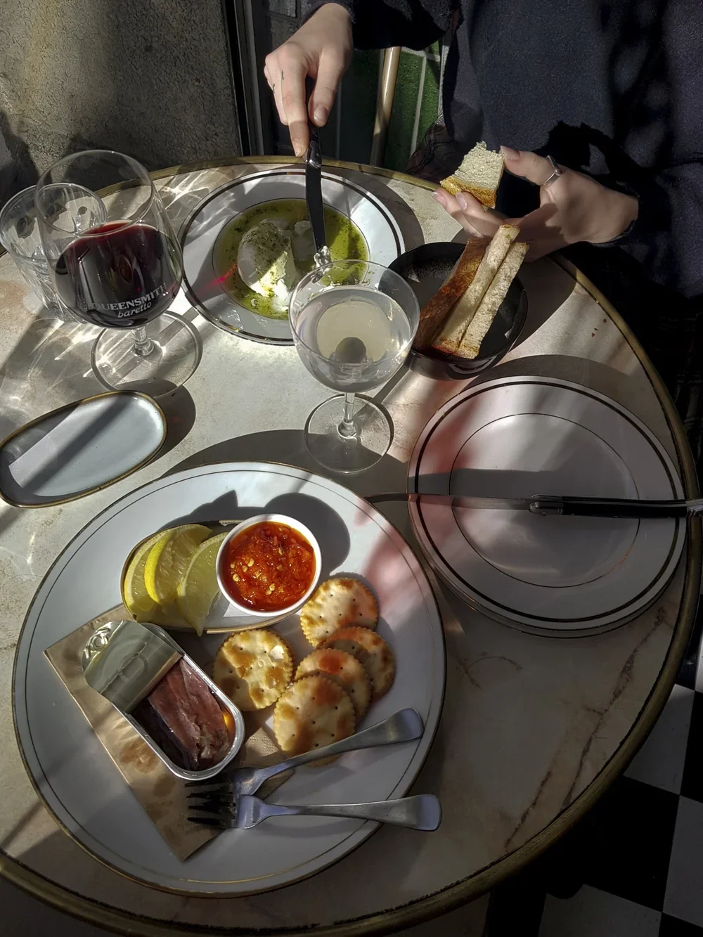 Table with a plate of sliced lemon, red chili sauce, and fried snacks, a container of cured meat, a fork and knife, glasses of red wine and white wine, a small dish of green olives, a bowl of green sauce, and a person holding bread and spreading some