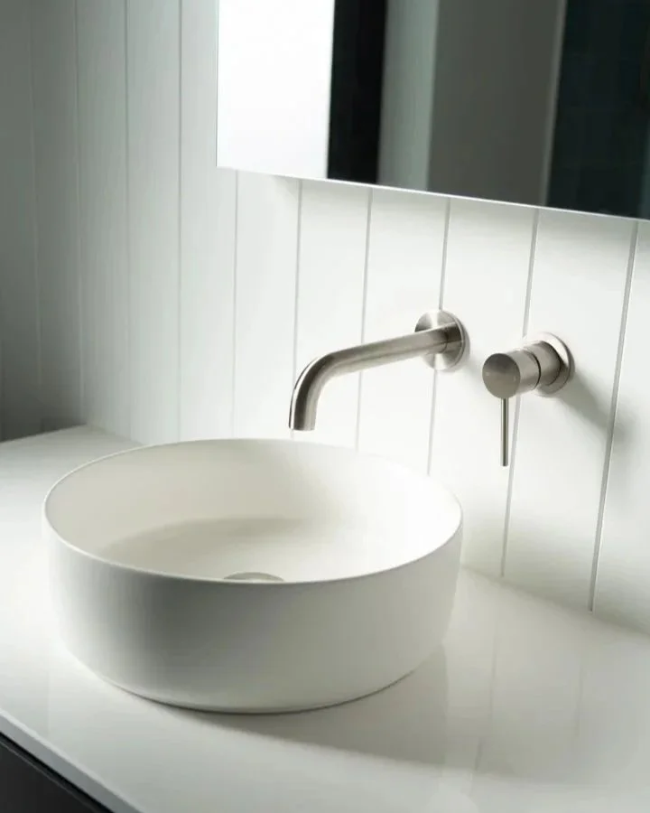 A modern bathroom sink with a round white vessel basin, wall-mounted stainless steel faucet, and a wall-mounted soap dispenser, set against a white tiled wall.
