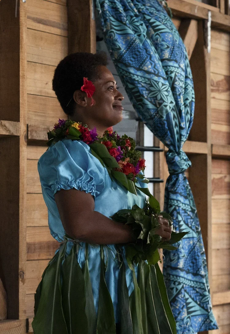 A woman with short dark hair, wearing a bright blue traditional dress and a floral lei, holding a bouquet of flowers, standing indoors with a wooden wall and a blue and black patterned curtain in the background.