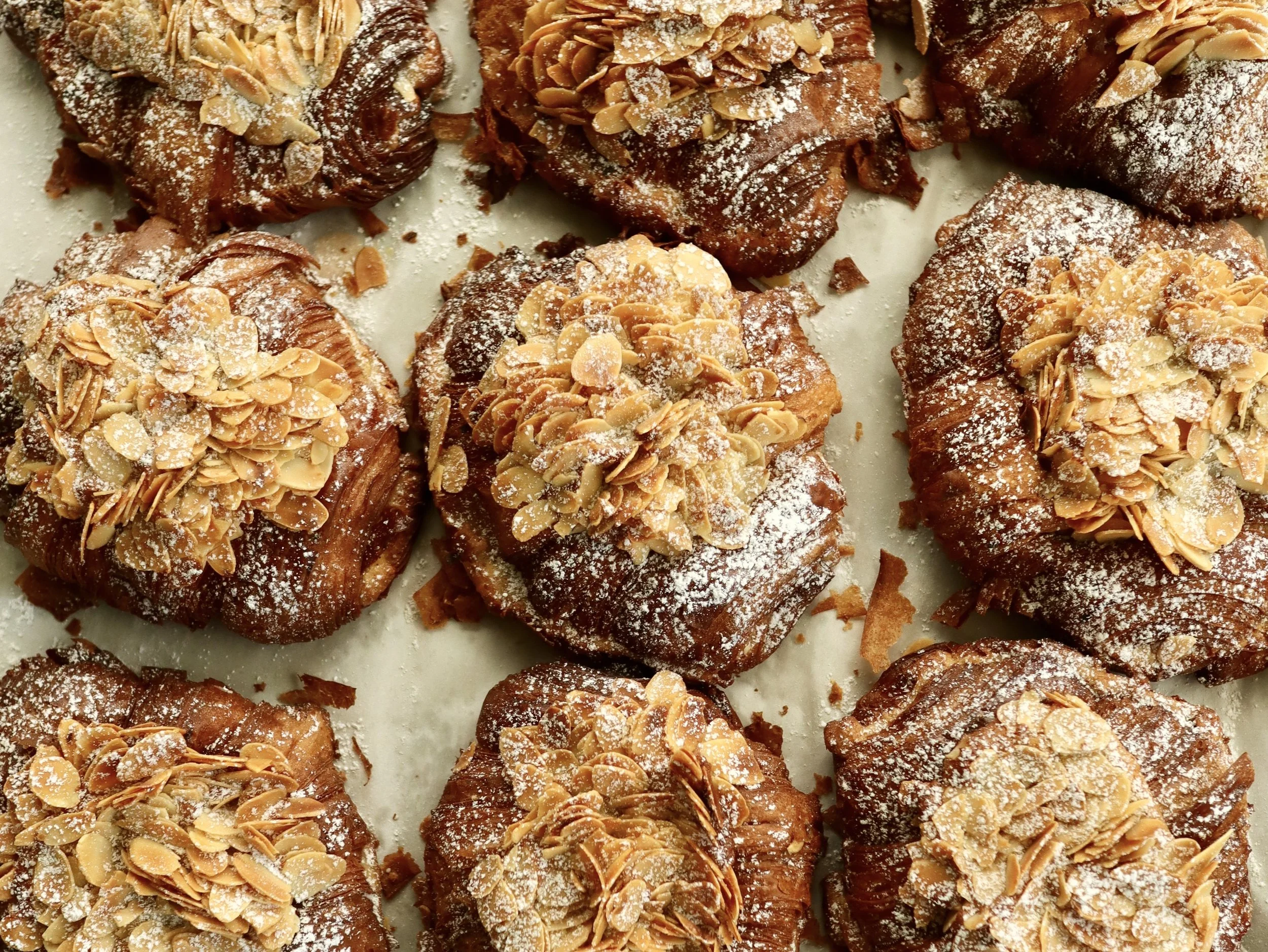 Close-up of several almond croissant pastries topped with sliced almonds and powdered sugar.