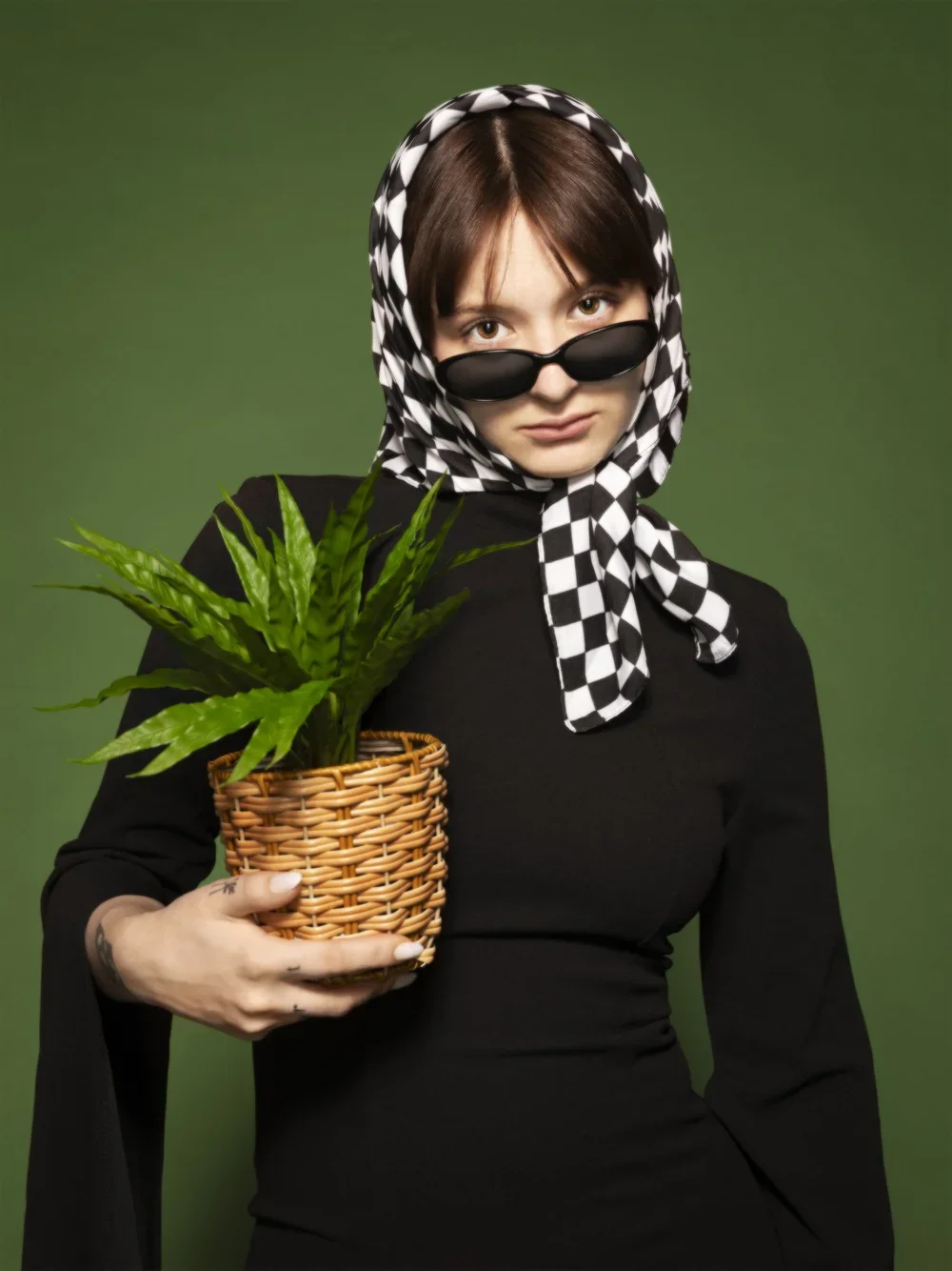 Woman with short brown hair wearing black sunglasses, a black and white checkered headscarf, and a black dress, holding a wicker basket with a potted green plant against a green background.