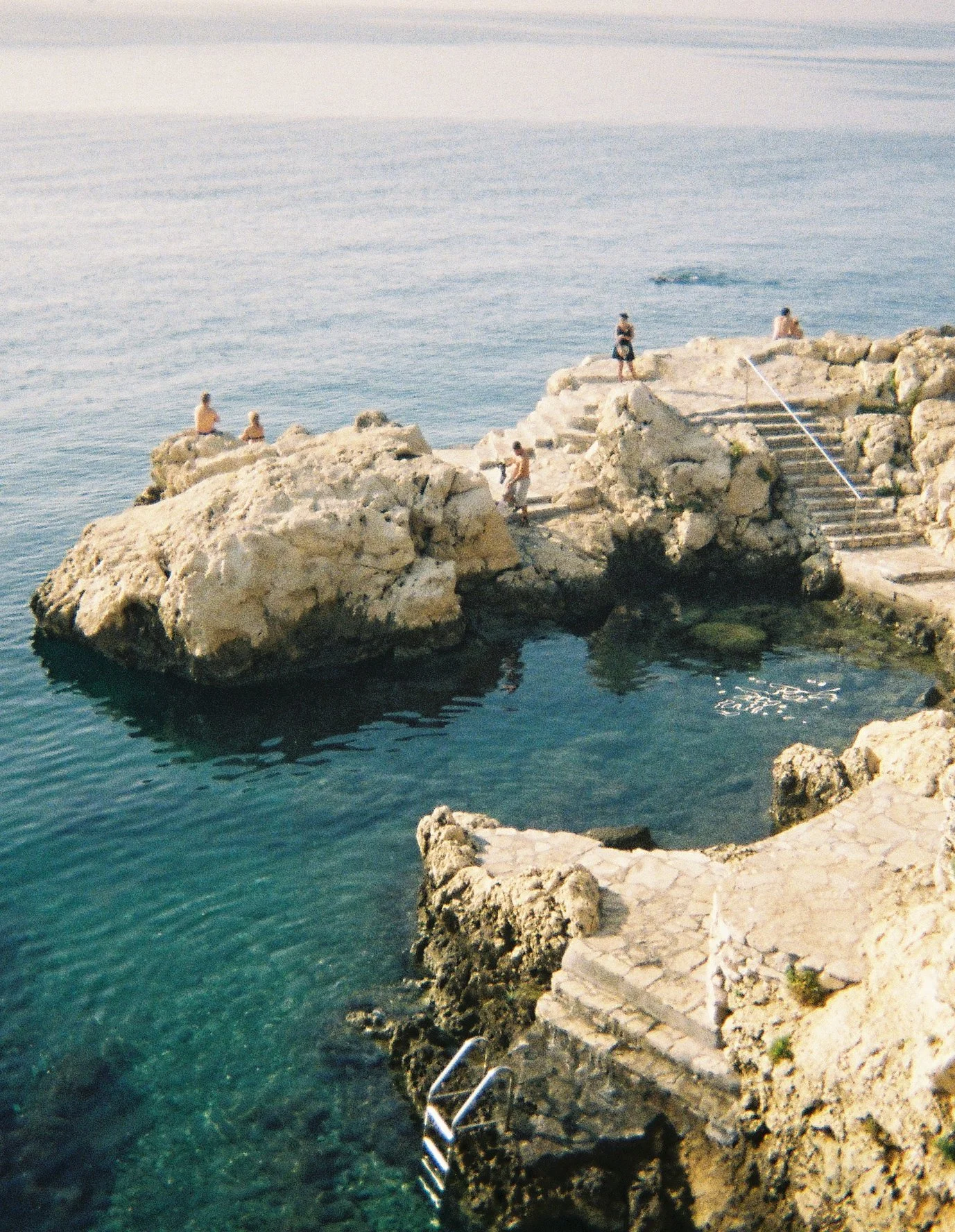 A rocky coastal area with stairs leading to the water, with five people relaxing and standing on the rocks near the water, and others sitting on the rocks.