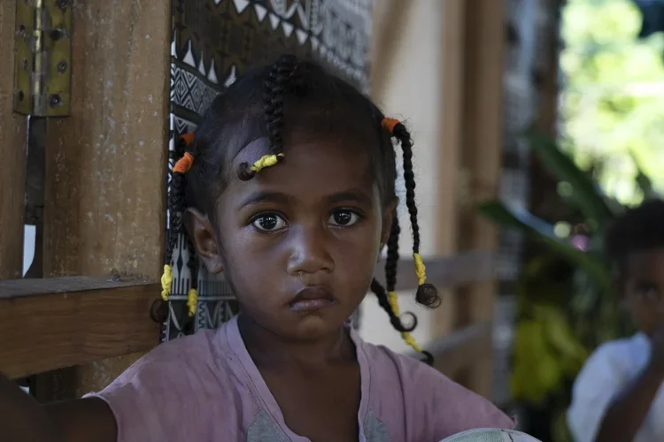 A young girl with braided hair decorated with colorful beads, sitting indoors near a wooden wall, looking directly at the camera with a serious expression.