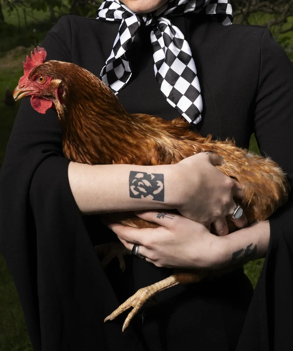 A person holding a brown chicken outdoors, wearing a black outfit and a black-and-white checkered scarf, with visible tattoos on their arms, including a rose and a square-shaped design.