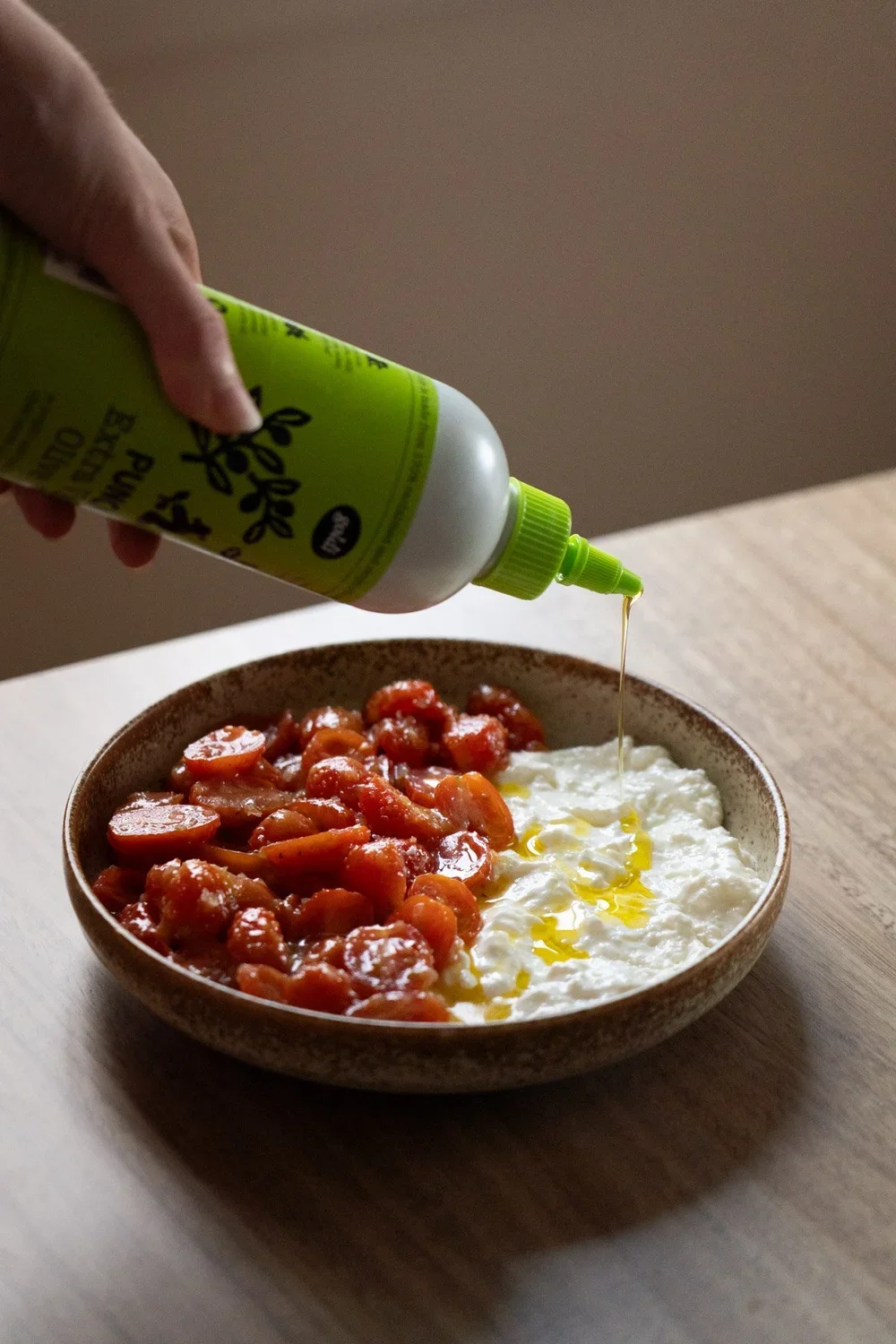 A bowl of cheese topped with tomatoes, with a person pouring oil from a green bottle.