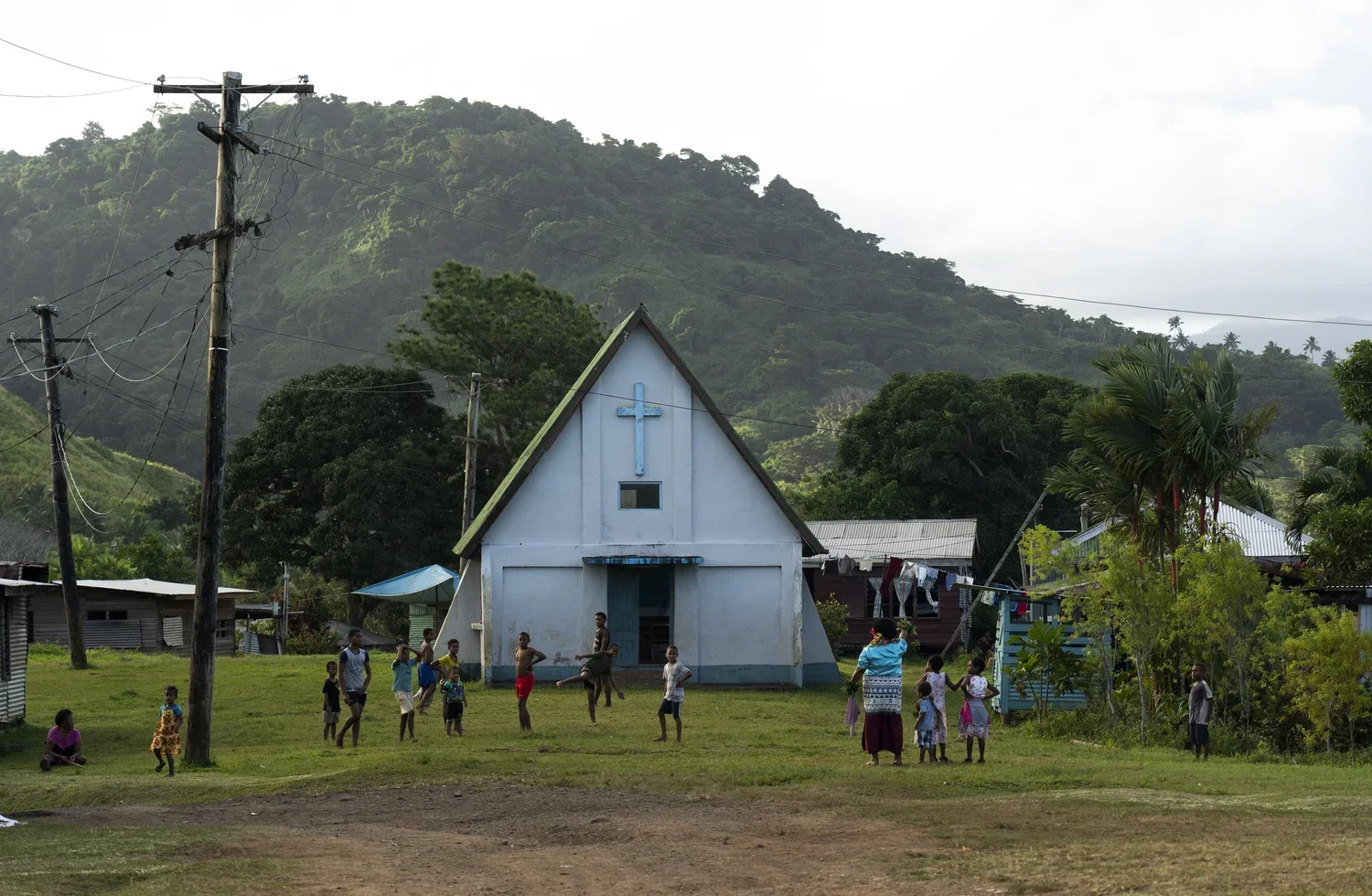 Children and women playing and standing in front of a small white church with a blue cross on the front, surrounded by green trees and hills in the background.