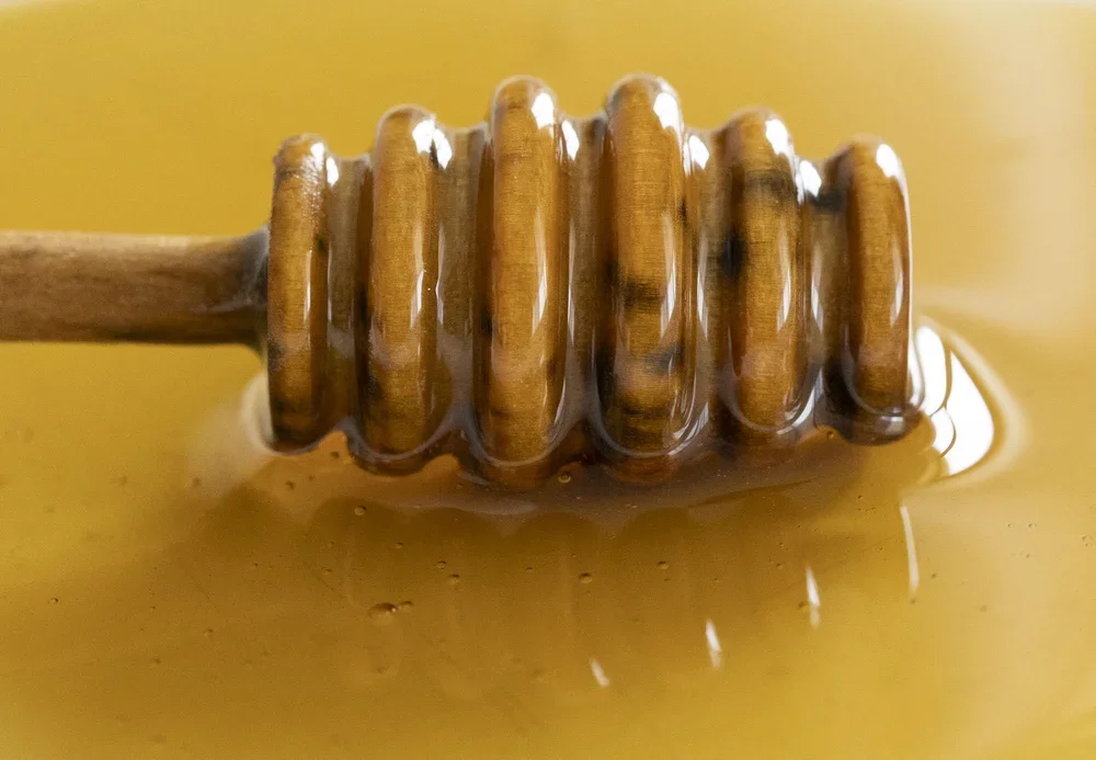 A wooden honey dipper coated in honey, with honey dripping off and pooling below on a yellow background.
