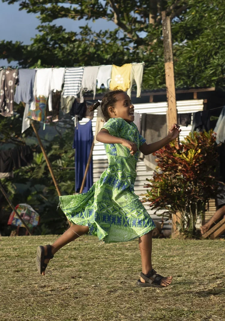 A young girl running outdoors in a grassy area, wearing a flowing green patterned dress and sandals, with laundry hanging on a line and trees in the background.