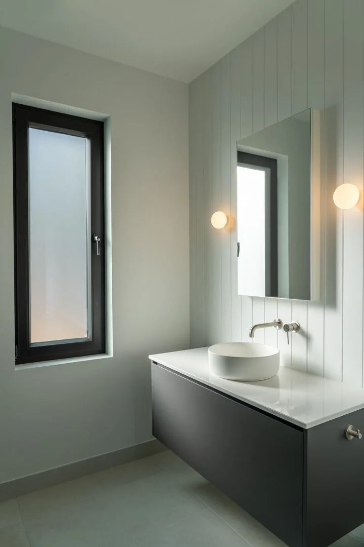 Modern bathroom with a white vessel sink on a black vanity, a large mirror, two wall-mounted spherical lights, and two tall narrow windows with frosted glass.
