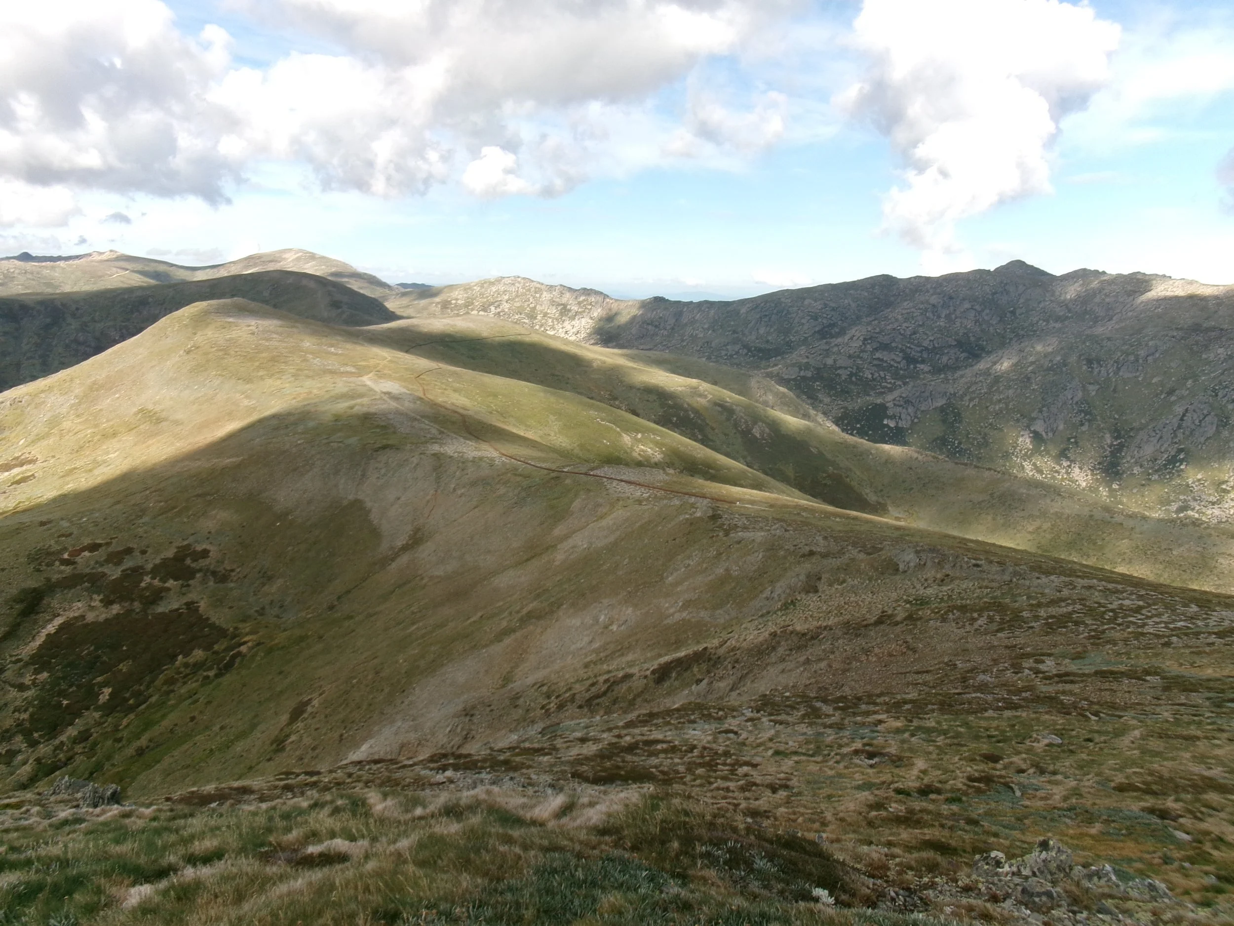 Scenic view of rolling green mountains with a narrow trail winding along the ridges under partly cloudy sky.