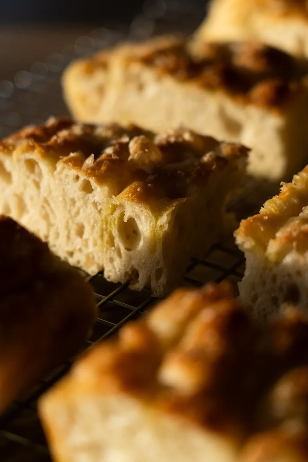 Close-up of a piece of focaccia bread on a wire rack, showing its airy interior and golden, rosemary-topped crust.