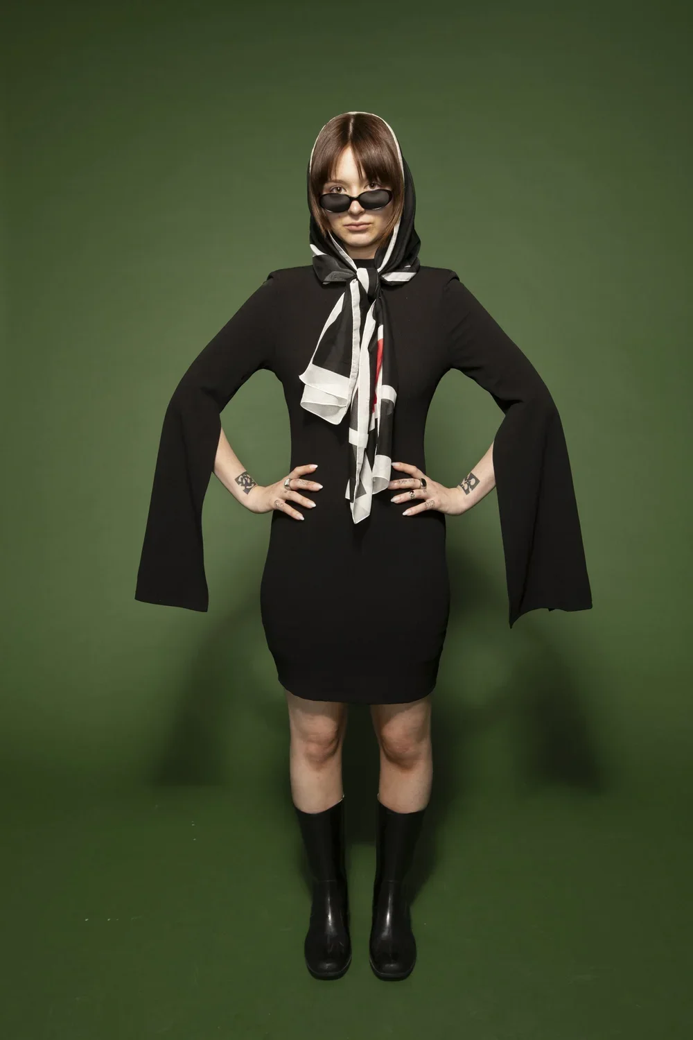 A woman with short brown hair wearing black sunglasses, a black dress with wide sleeves, a patterned scarf, and black boots, standing with her hands on her hips against a green background.