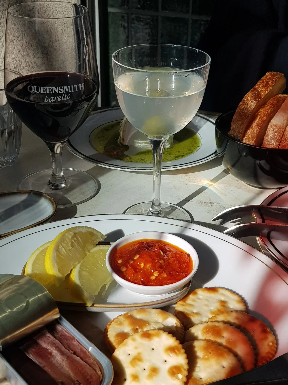 A table set with glasses of red and white wine, a dish of sliced bread, a small bowl of tomato-based dipping sauce, lemon wedges, crackers, and a container of sliced cured meat.