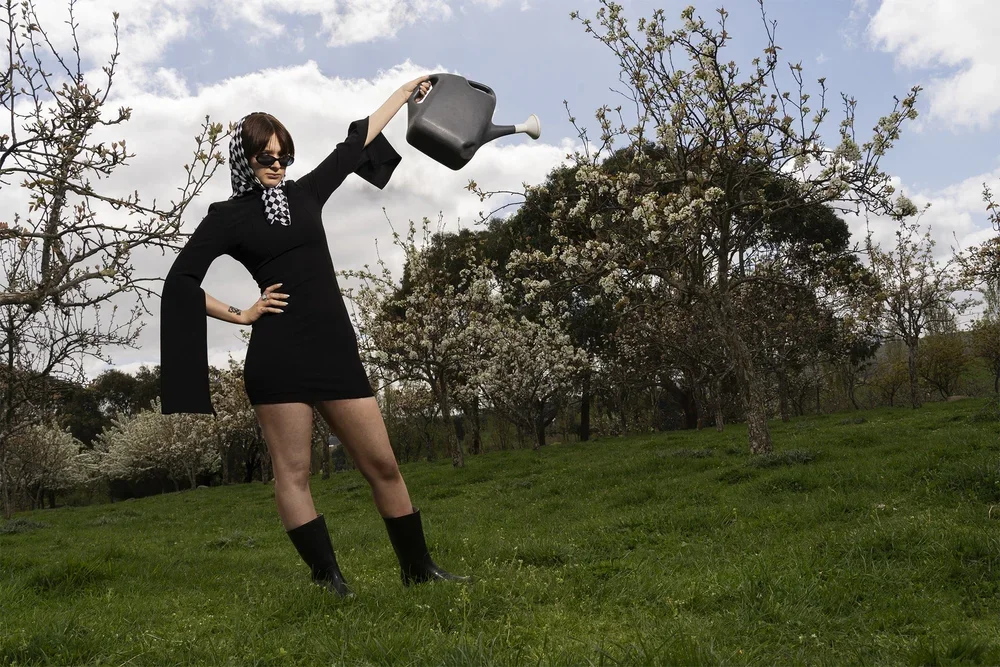 A woman in a black dress with bell sleeves, checkered scarf, and black boots, stands in a grassy field watering plants with a large gray watering can, surrounded by blossoming trees under a cloudy sky.