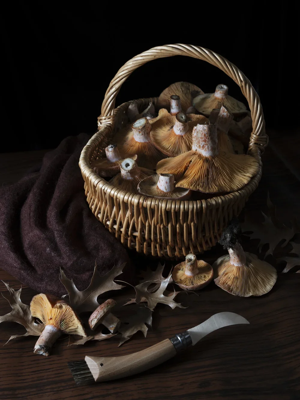 Basket of wild mushrooms with dried leaves and a small knife on a dark wooden surface.