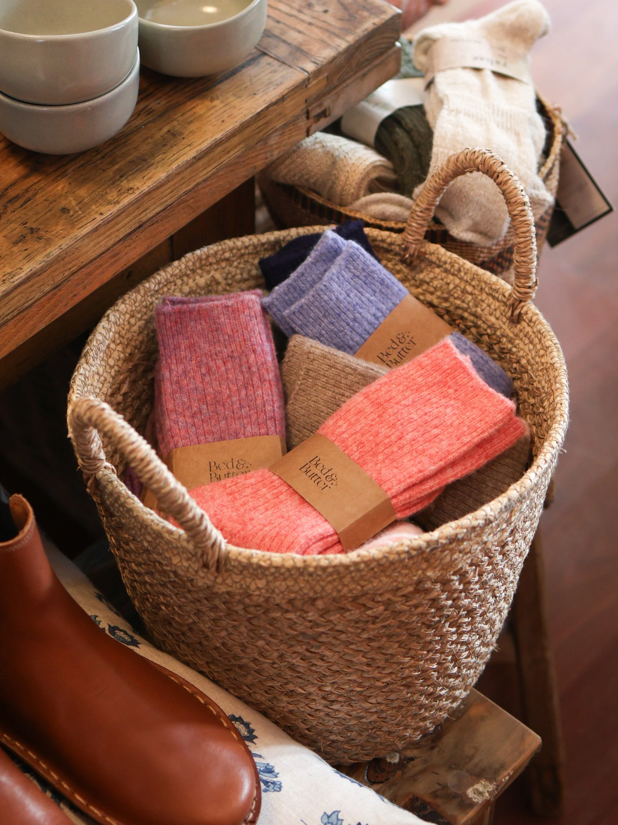 A woven basket filled with assorted folded colorful socks with labels, set on a wooden surface next to a pair of brown boots and a wooden table with stacked white bowls.