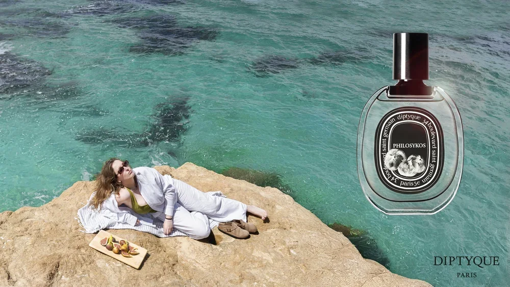 A woman with blonde hair in a white outfit relaxing on a rock by the sea with a tray of peaches, looking up at a large perfume bottle labeled 'PHILOSYKOS' floating in the water.