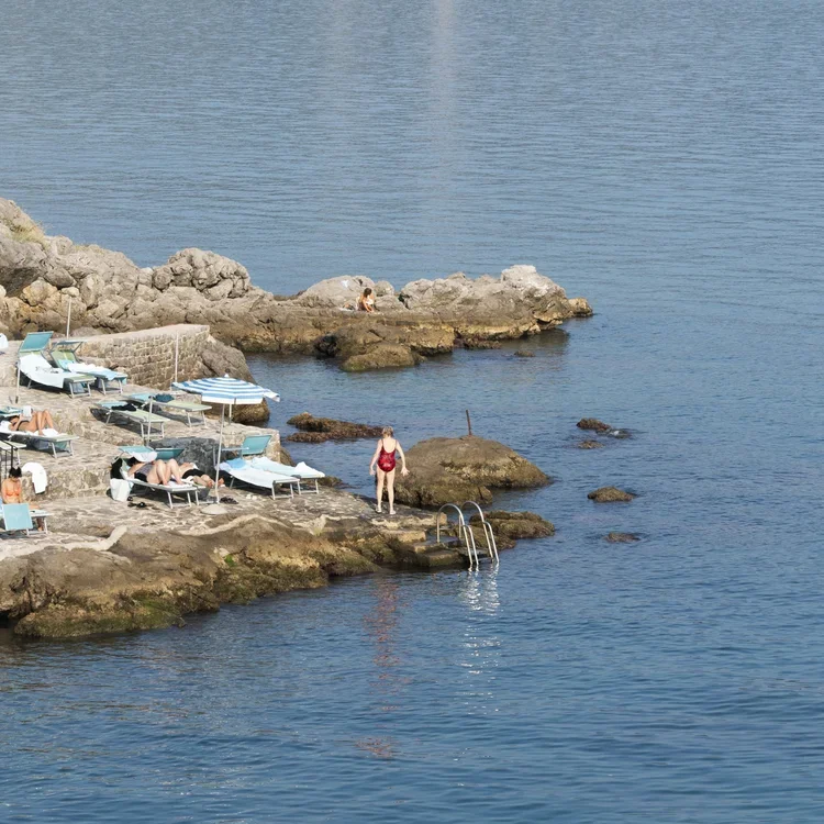 People relaxing on rocky ledges by a body of water, some on lounge chairs with umbrellas, others sitting on rocks, with a ladder leading into the water.