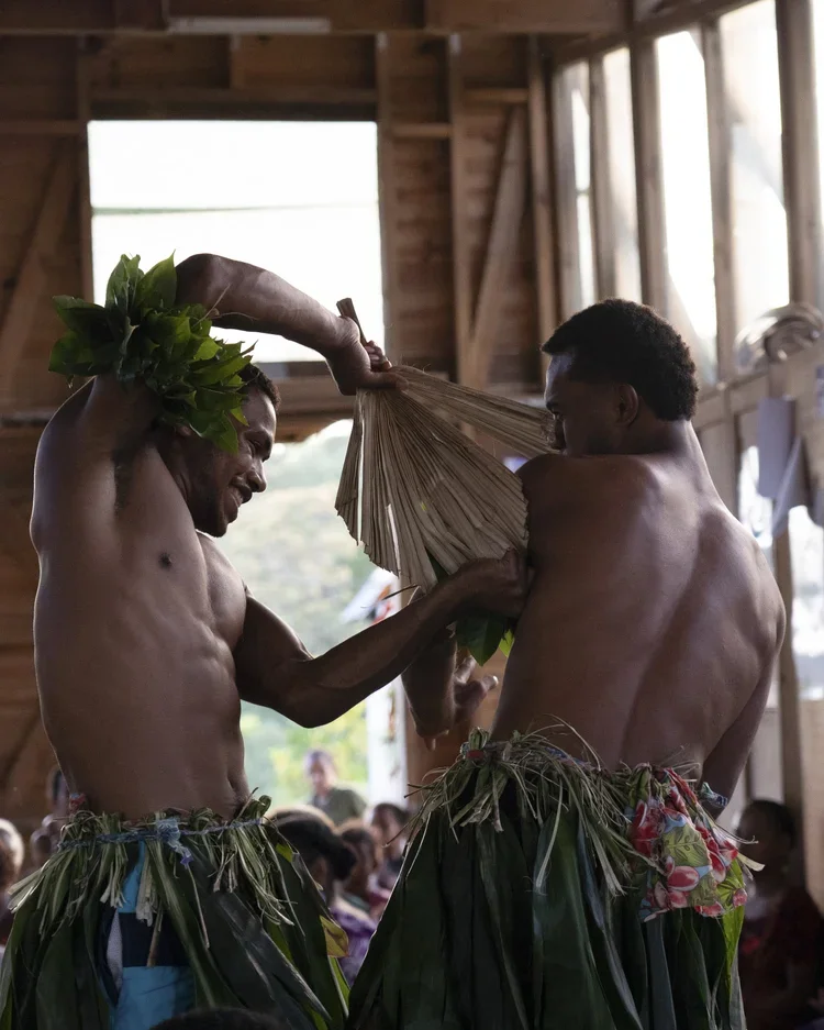 Two shirtless men wearing traditional grass skirts are dancing and holding a leafy object inside a wooden building with large windows, with an audience watching in the background.