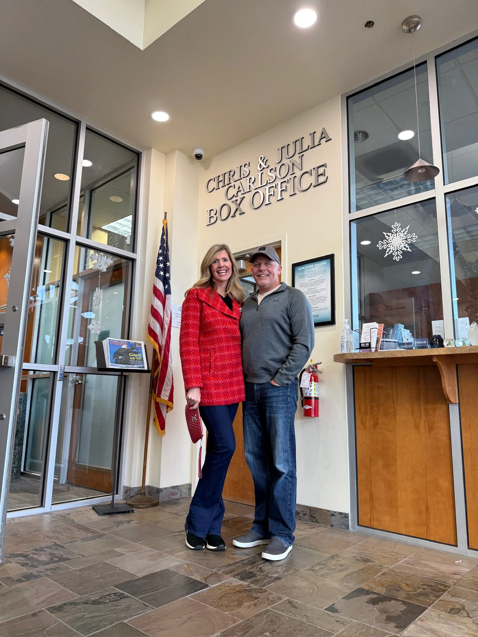 A woman in a red coat and a man in a gray jacket standing inside a building in front of the Chris & Julia Carlson Box Office sign. An American flag is displayed nearby, and frosted glass windows with snowflake decorations are visible.