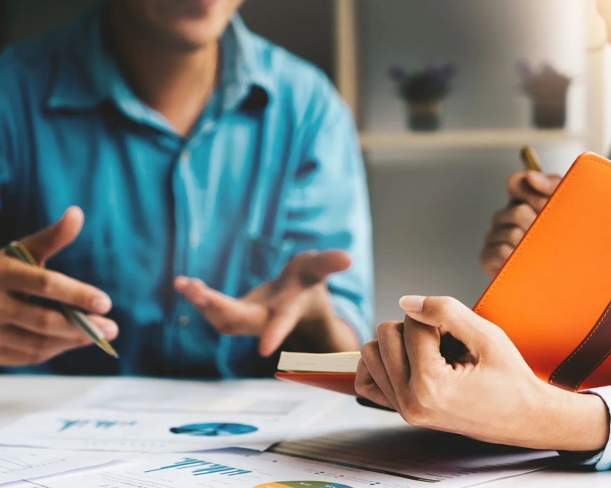 Two people having a discussion at a table, one person holding an orange notebook and the other holding a pen, with documents and charts on the table.