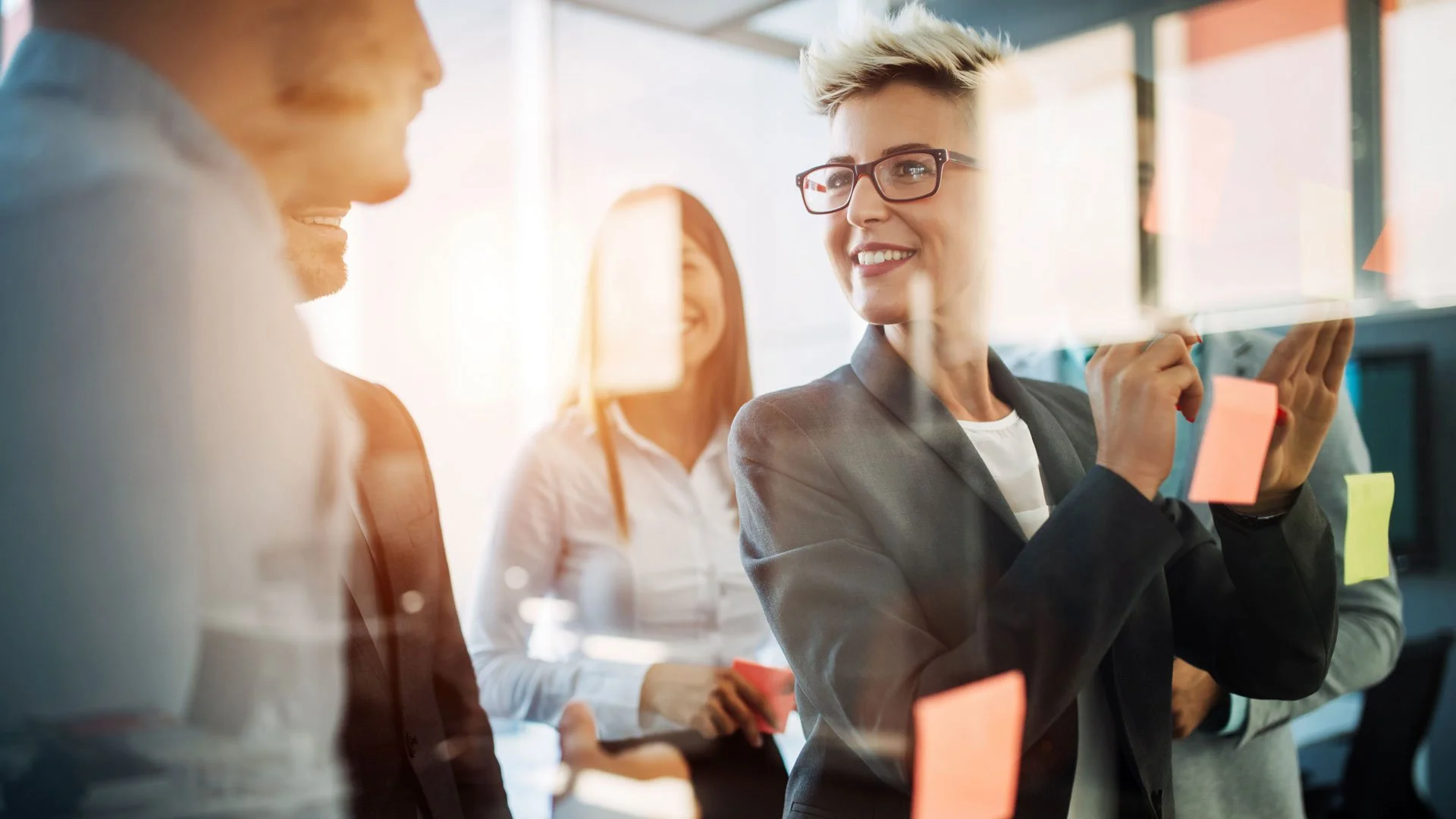 A group of professionals collaborating in a modern office. One woman with short blonde hair and glasses is smiling and placing a sticky note on a glass wall, while others observe and participate in the discussion.