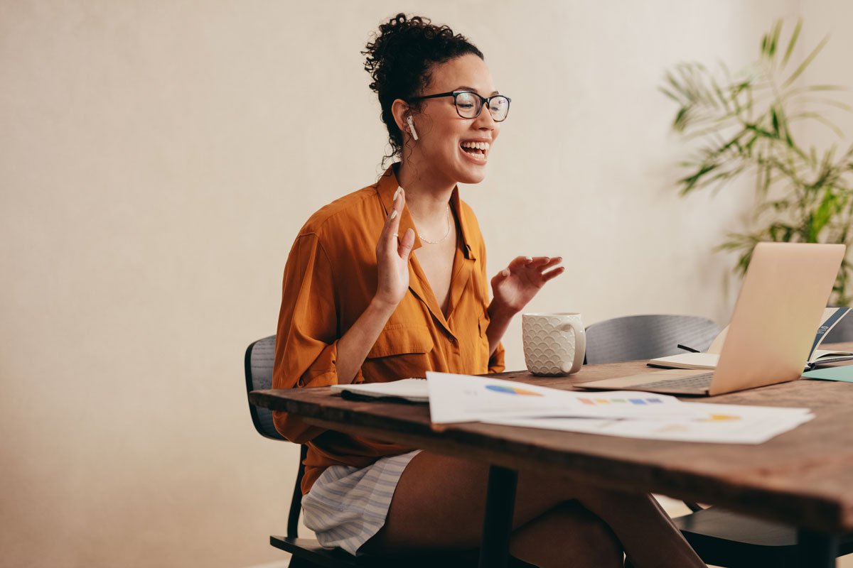 Woman smiling and talking during a video call at her desk with laptop, papers, mug, and a plant in the background.