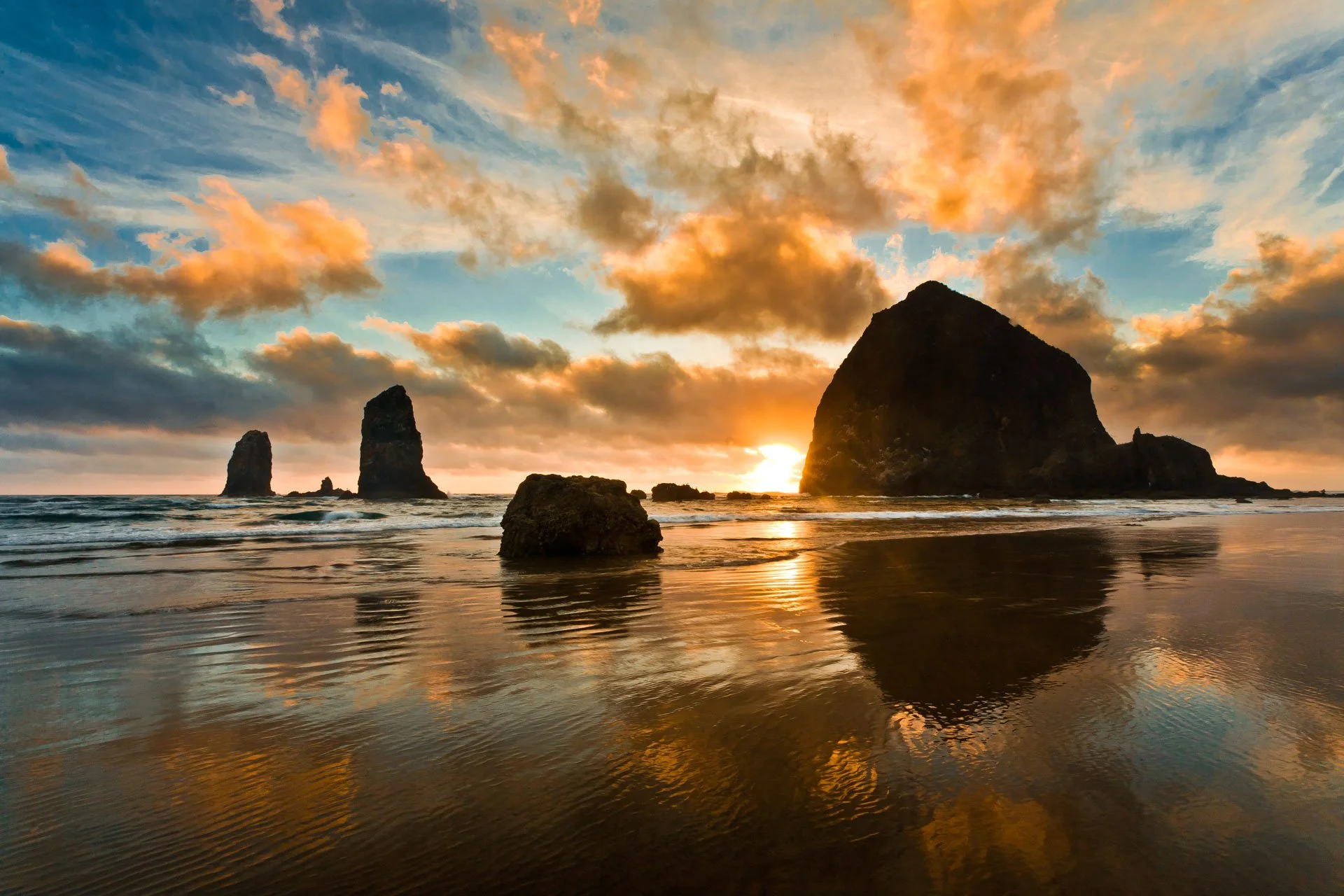 A breathtaking sunset over the ocean with large rock formations rising from the water, some partly submerged, and colorful clouds in the sky reflecting on the wet sand.
