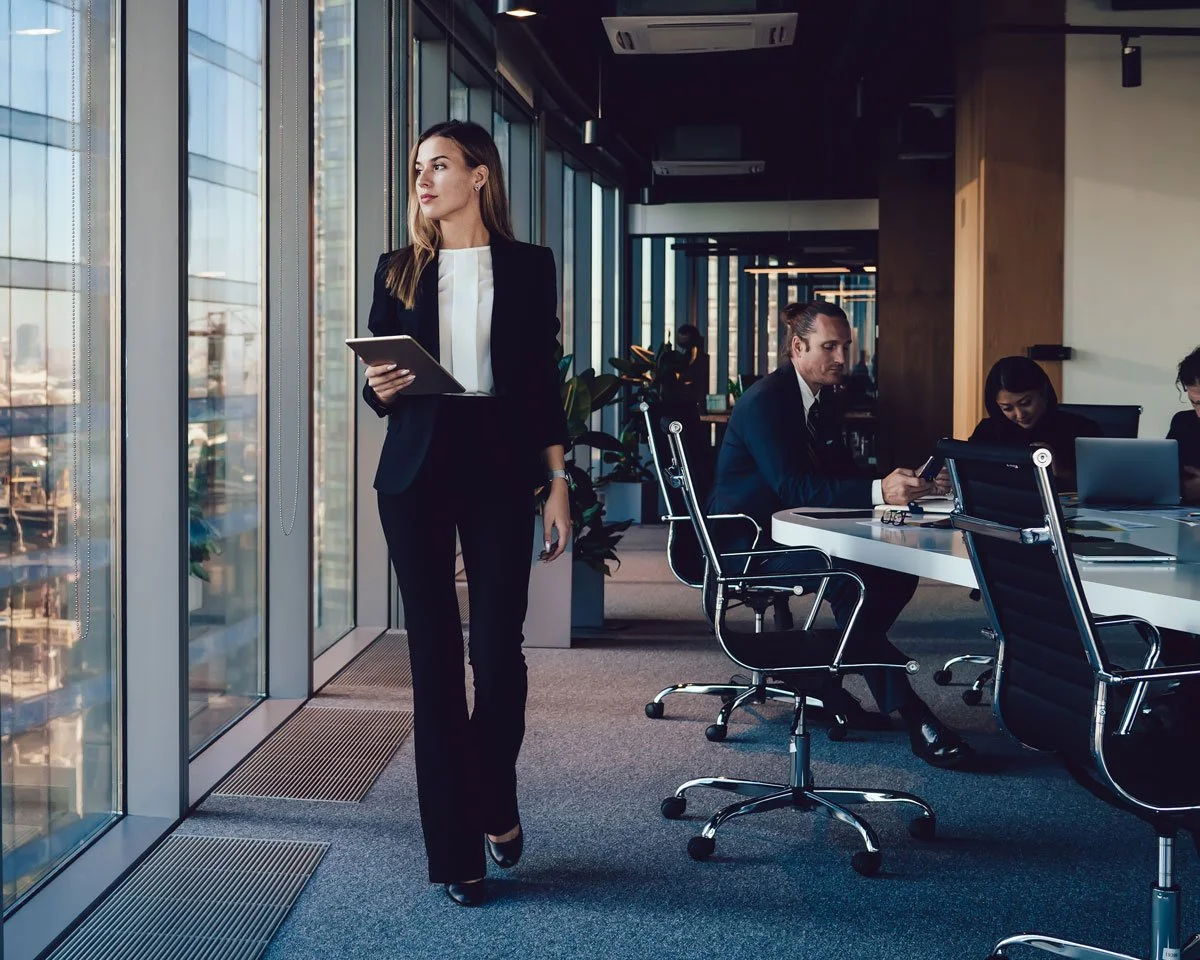 A woman in a business suit holding a tablet walking in a modern office with large windows, while colleagues work at a table in the background.