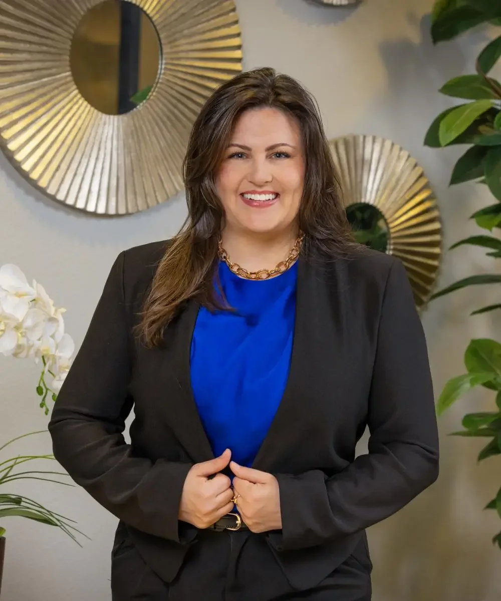 A smiling woman with brown hair, wearing a black blazer, blue shirt, and gold necklace, standing in front of a wall with circular gold mirrors and a green plant.