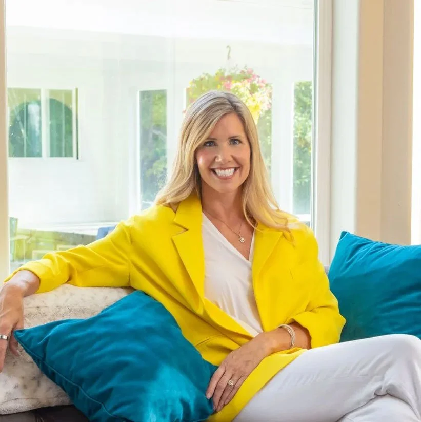 A woman with blonde hair smiling, wearing a yellow blazer and white clothes, sitting on a couch with blue cushions in a bright room with a large window.