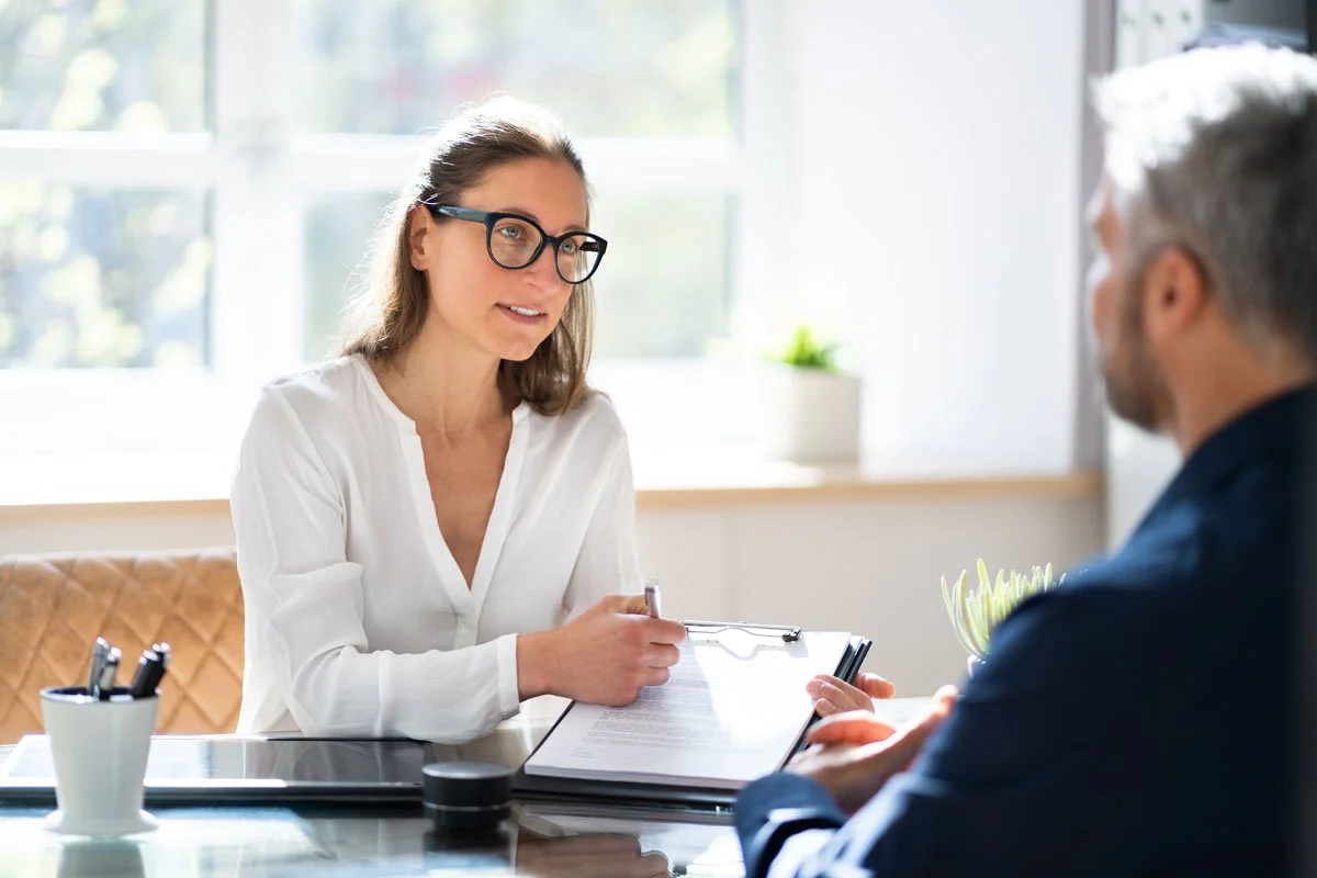 A woman with glasses and a white blouse sitting at a desk during a meeting with a man.