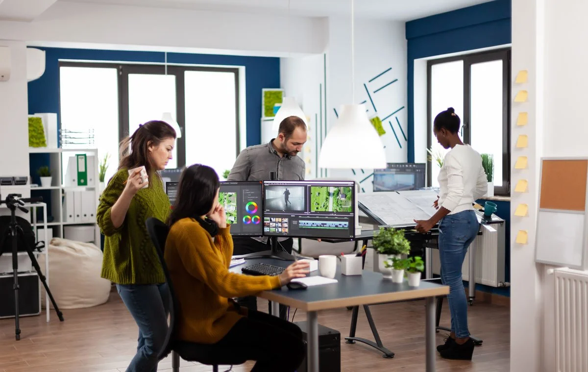 Four people working in a modern office space with computer monitors, a tripod, and various office supplies, during daylight.