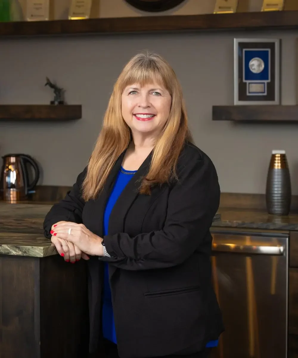 A woman with long reddish-blonde hair and red lipstick, wearing a black blazer over a blue top, standing in a modern kitchen with a wooden counter, a metallic kettle, and framed awards or certificates on the wall behind her.