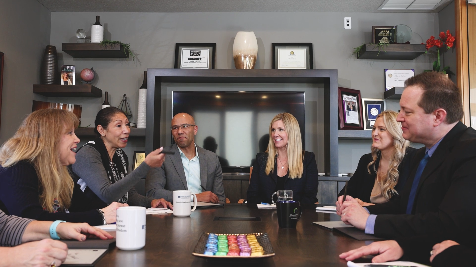A diverse group of six professionals sitting around a conference table in a modern office, engaged in discussion and smiling. The table has a tray of colorful candies and some coffee mugs.